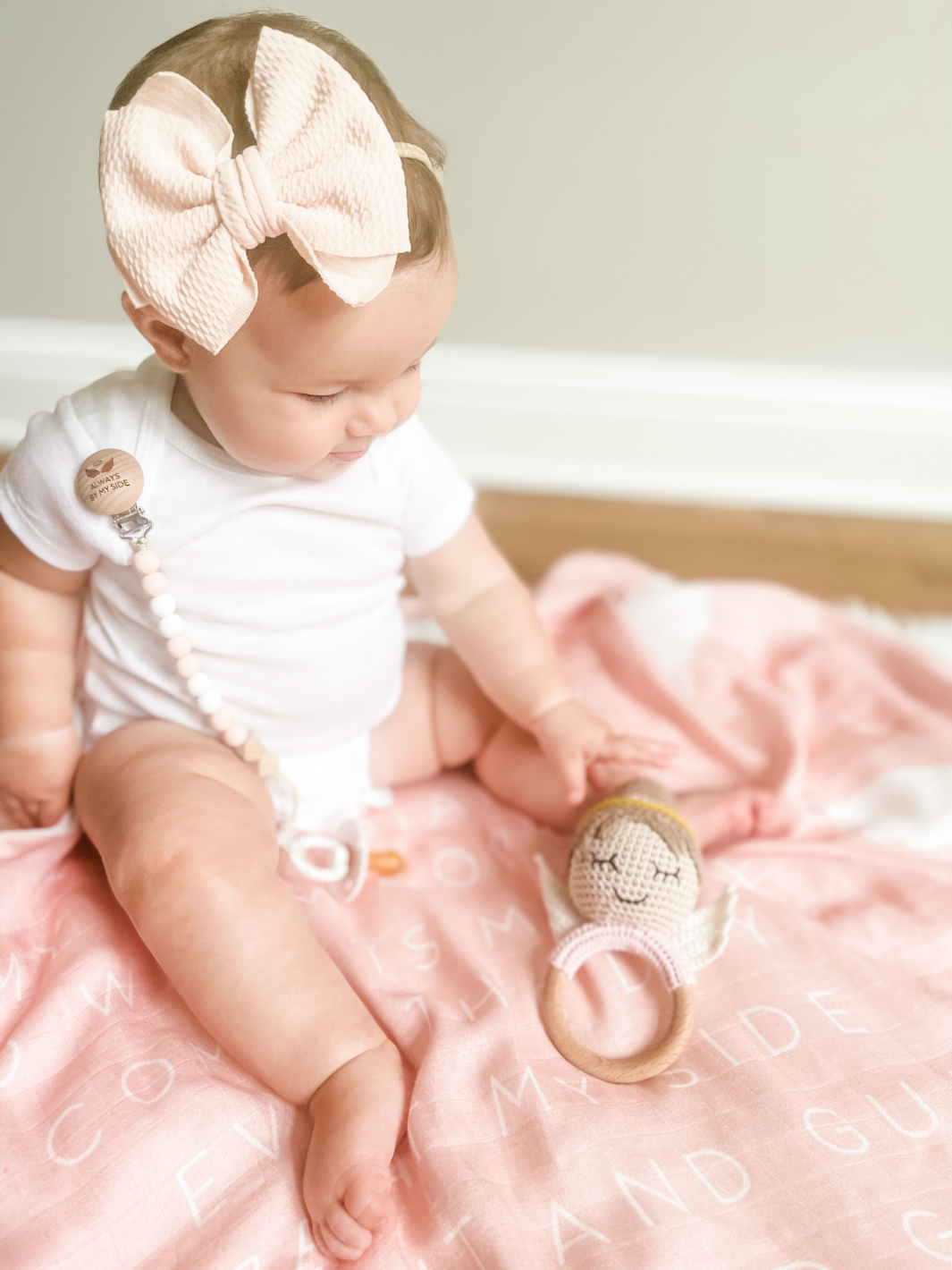 Baby sitting on a pink blanket wearing a white onesie and large bow headband, holding a teething ring.