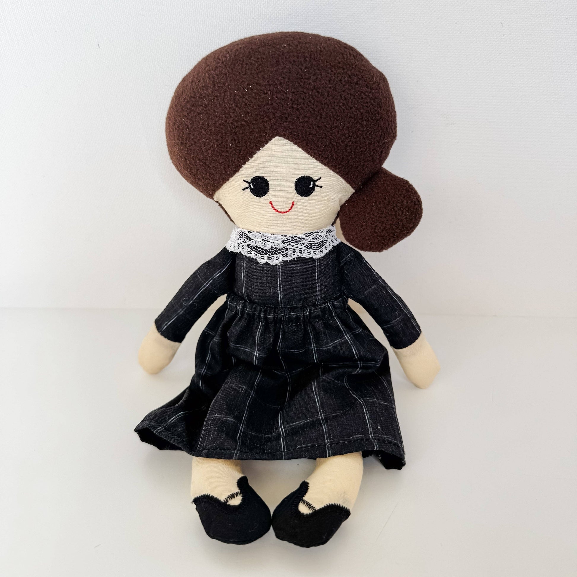 Plush doll with brown hair styled in pigtails, wearing a black dress with white lace collar on a light gray background.
