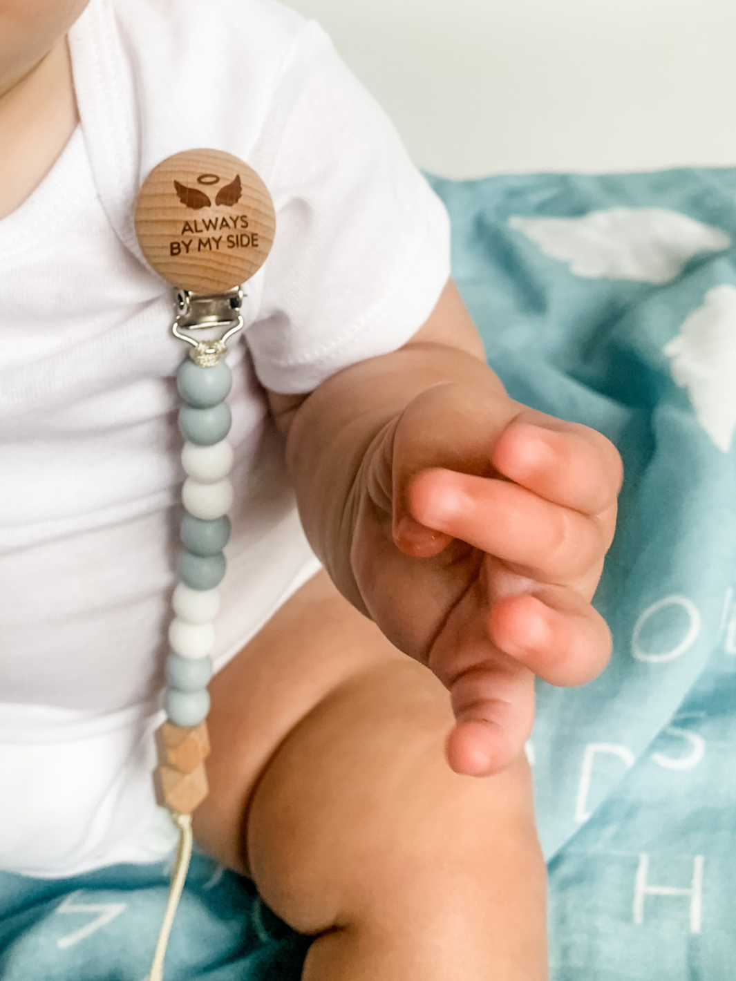 Baby holding a pacifier clip with wooden badge and beads, against a light blue blanket.