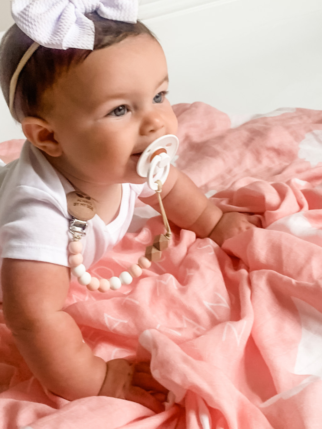 Baby in a pink dress with a white bow and pacifier, sitting on a pink blanket.