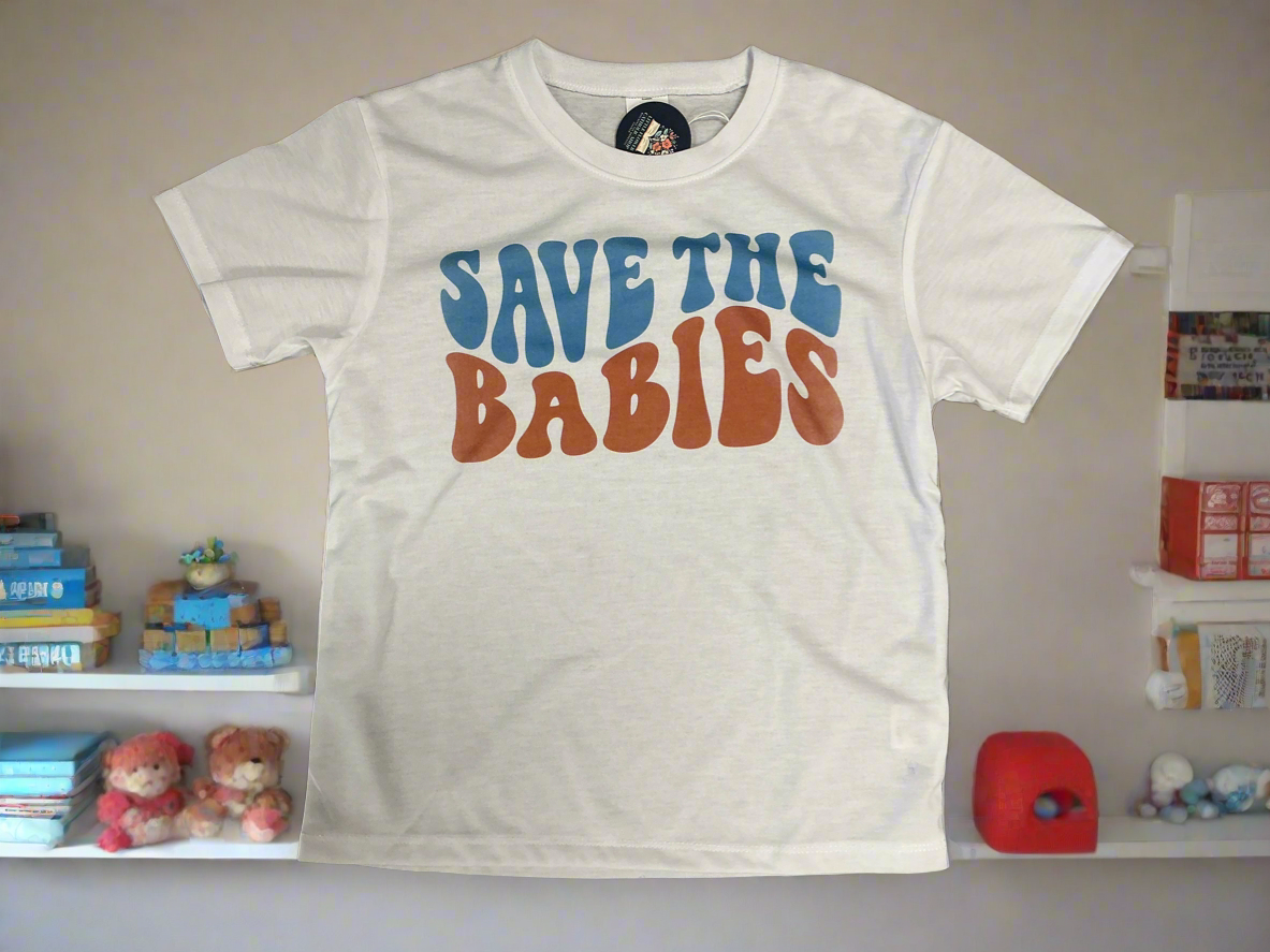 White t-shirt with 'SAVE THE BABIES' text in colorful letters on a shelf with toys.