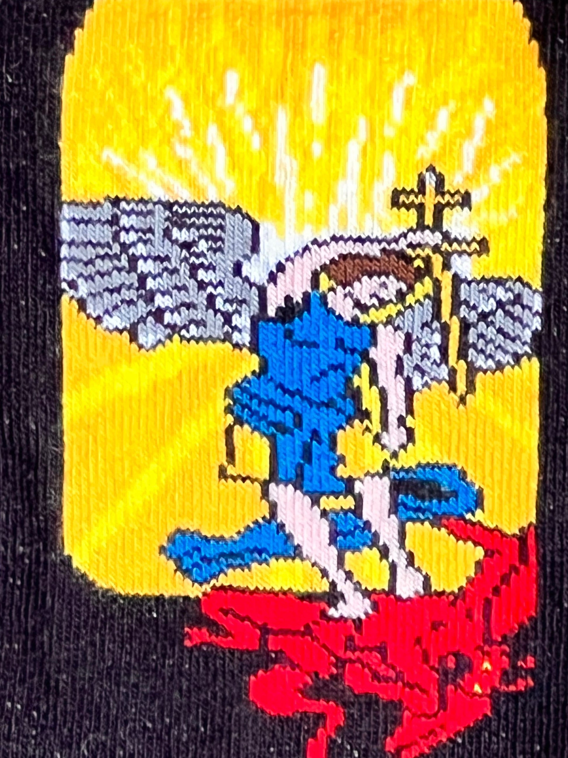 Embroidered design on fabric with a central figure holding a cross, surrounded by wings and flames.