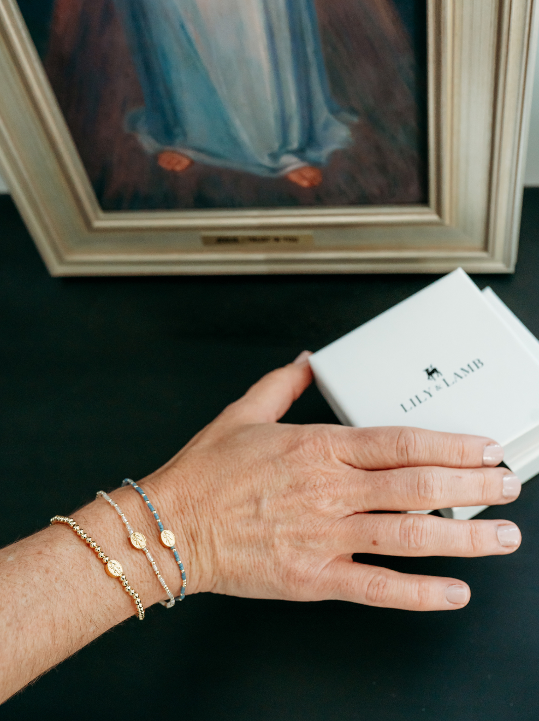 Hand with gold bracelets holding a white box labeled 'Jewelryland' in front of a framed painting.