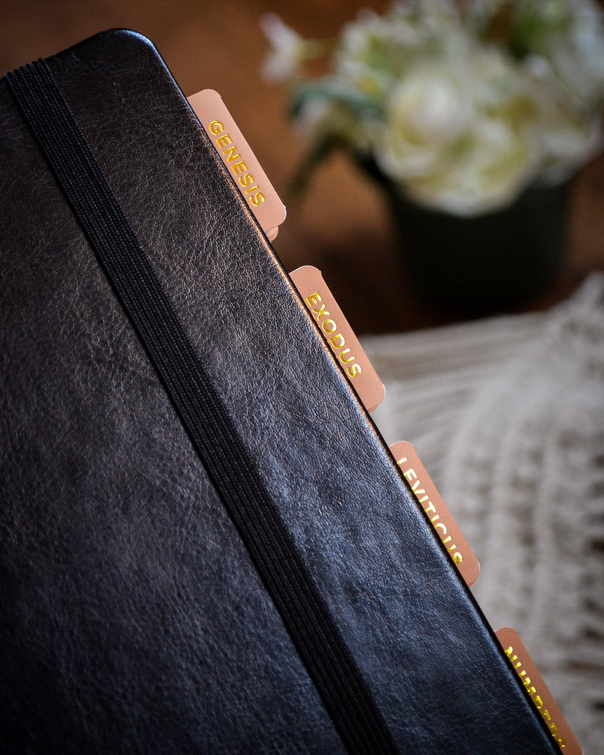 Black leather-bound book with gold lettered tabs on a wooden surface with flowers in the background