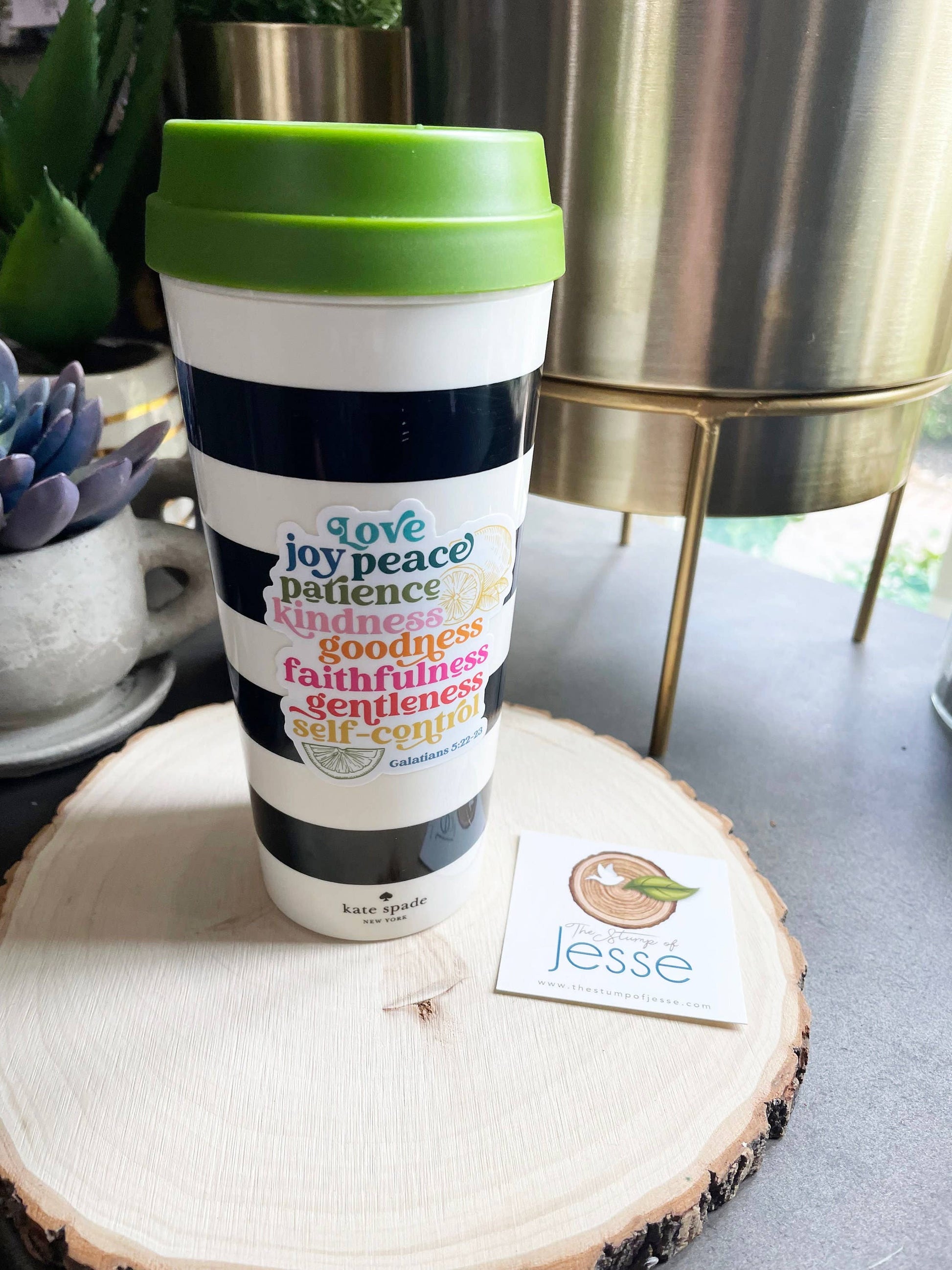 Travel mug with fruits of the Holy Spirit on a wooden coaster with a plant and decorative item in the background. 