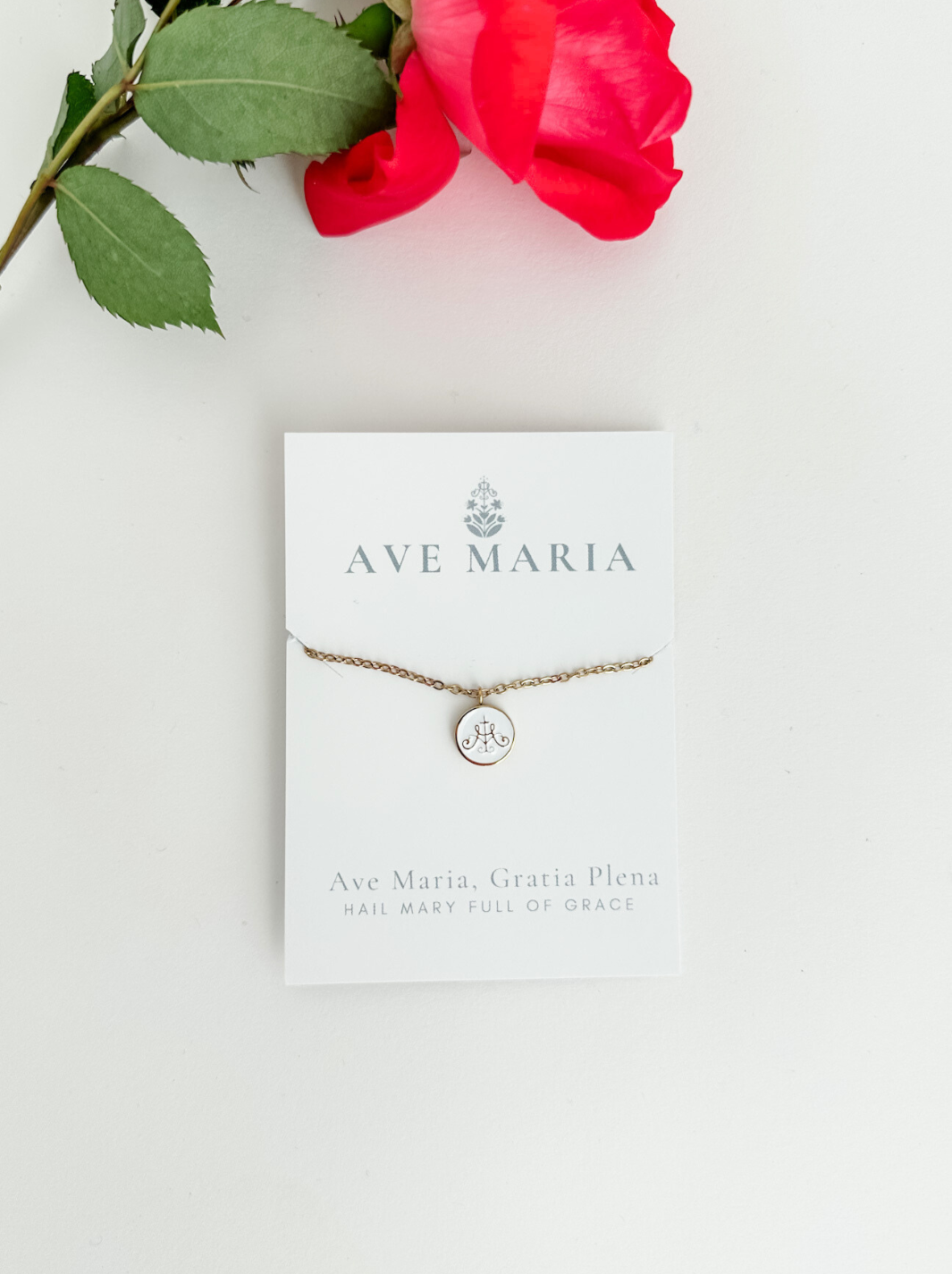 Ava Maria symbol on white background with gold border attached to golden chain with rose in background 