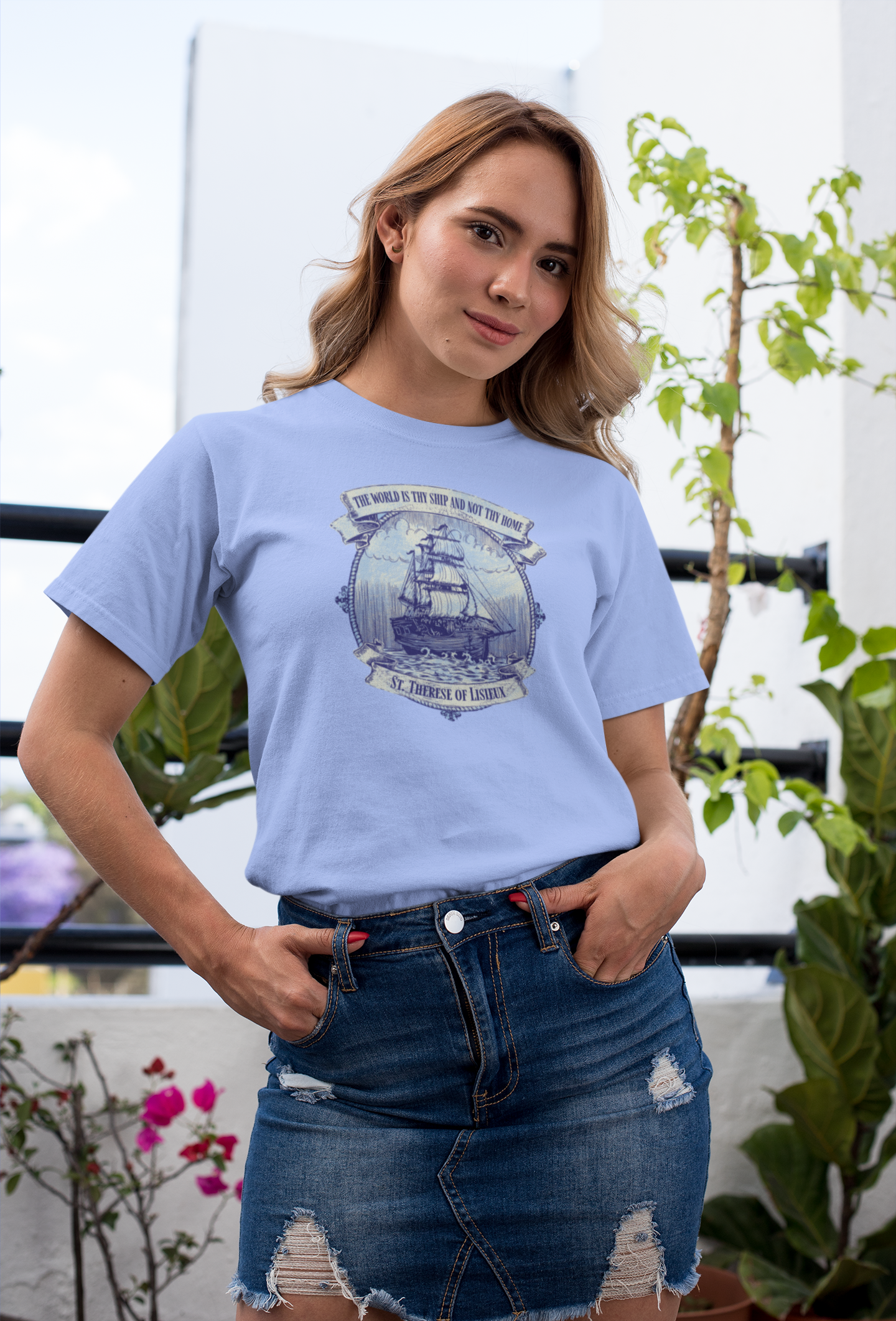 Woman wearing a light blue t-shirt with a graphic design, standing outdoors with plants in the background.