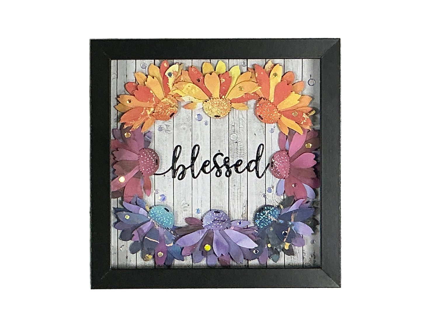 Framed decorative wall art with layered paper flowers in orange, yellow, pink, purple, and blue around the word 'blessed' on a whitewashed wood background, shown in a black frame against a gray textured wall.