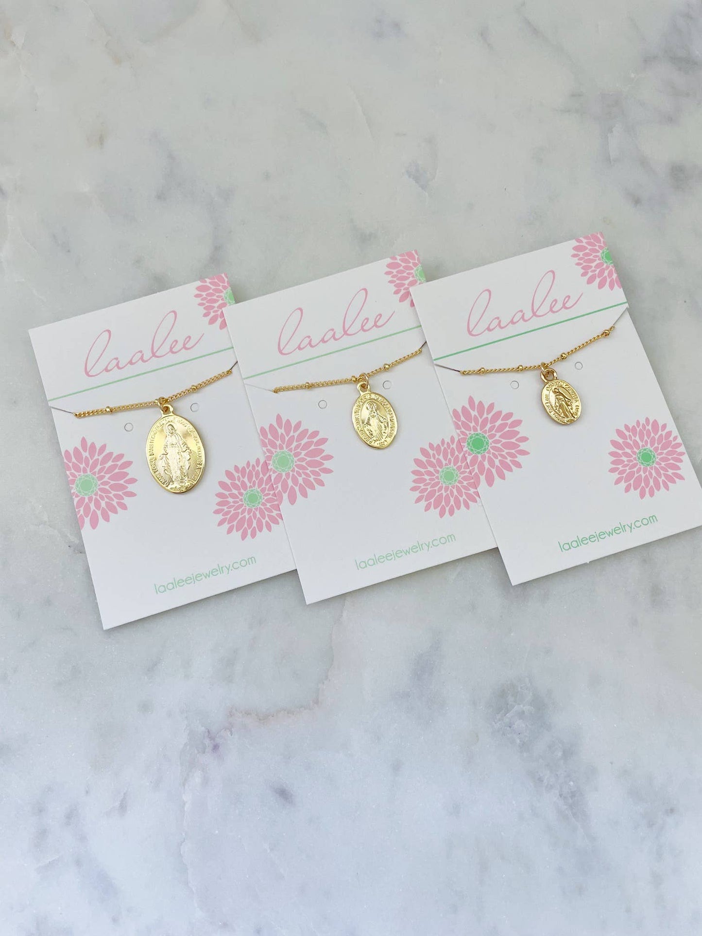 Three gold necklaces on 'Laalee' branded cards with pink floral designs on a marble surface.
