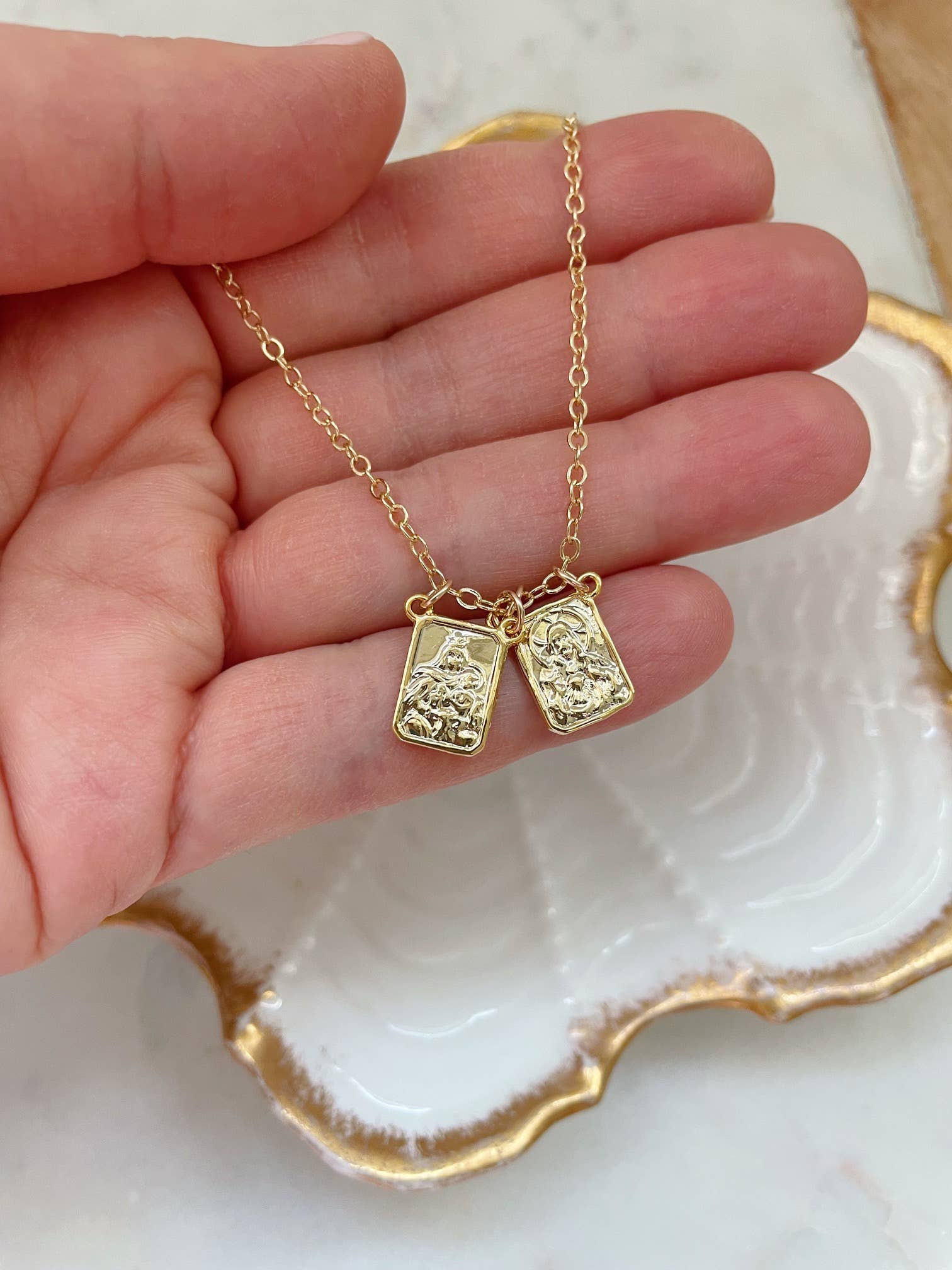 Gold necklace with two pendants held by a hand on a decorative plate.