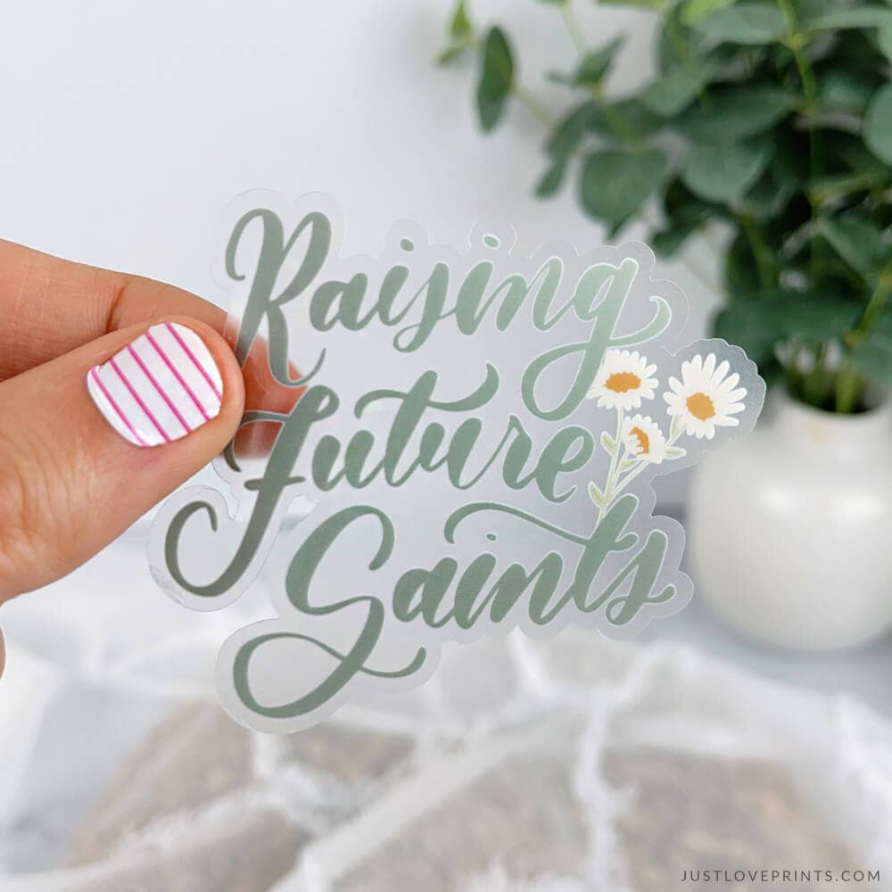 Clear acrylic sign with 'Raising future Saints' text held by a hand, with a plant in the background.