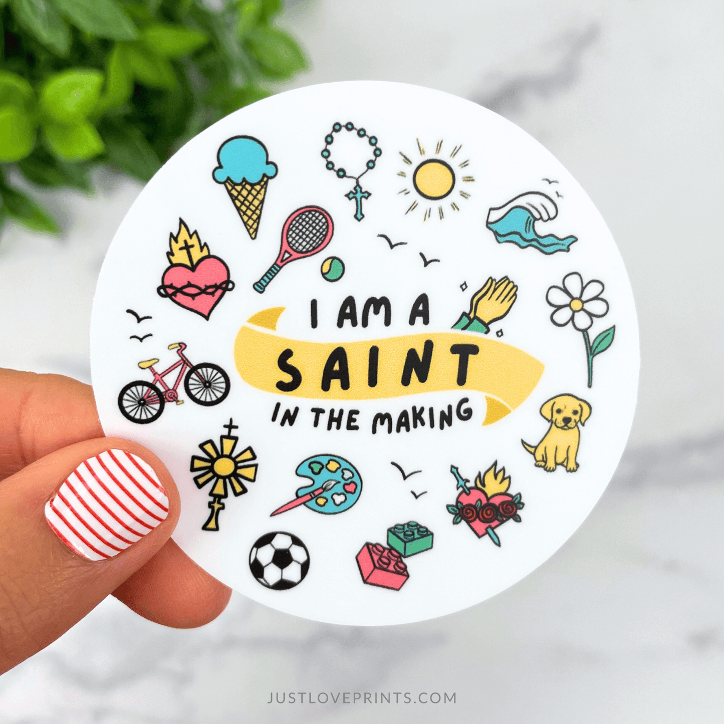 Circular sticker with illustrations and text 'I am a saint in the making' held by a hand.