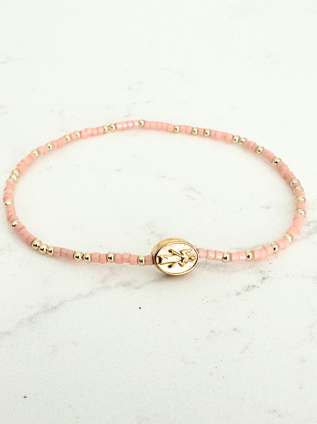 Pink beaded bracelet with a gold anchor charm on a marble surface