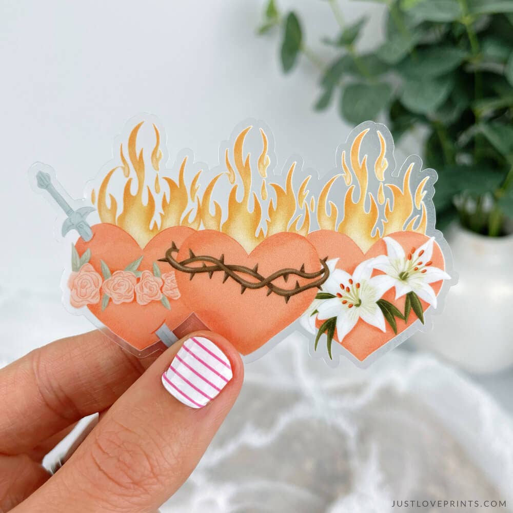 Heart-shaped sticker with flames, sword, and flowers held by a hand against a blurred natural background.