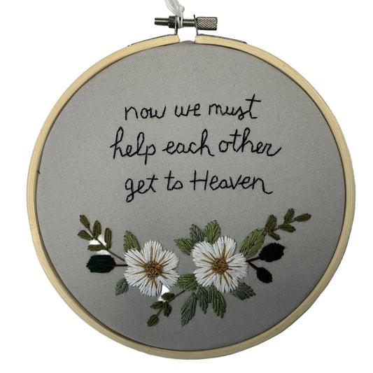 Embroidery hoop with text 'now we must help each other get to Heaven' and floral design on a gray background.