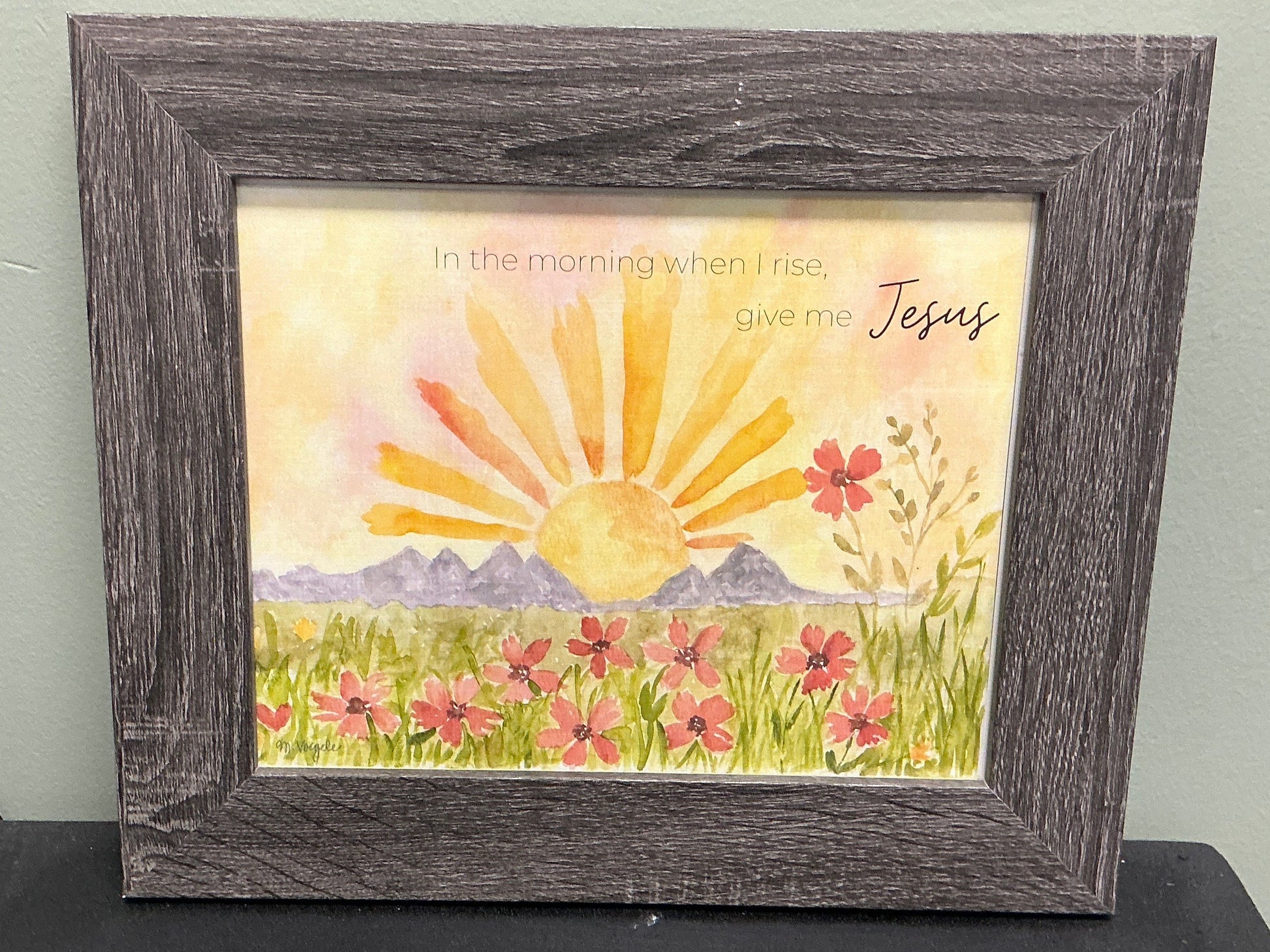 Framed artwork with a sun and mountain scene on a table against a plain wall.