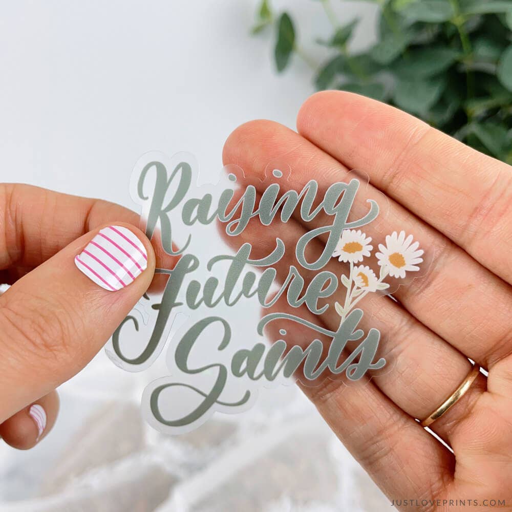 Hand holding a clear acrylic cutout with 'Raising future Saints' text and flowers, blurred greenery in the background.