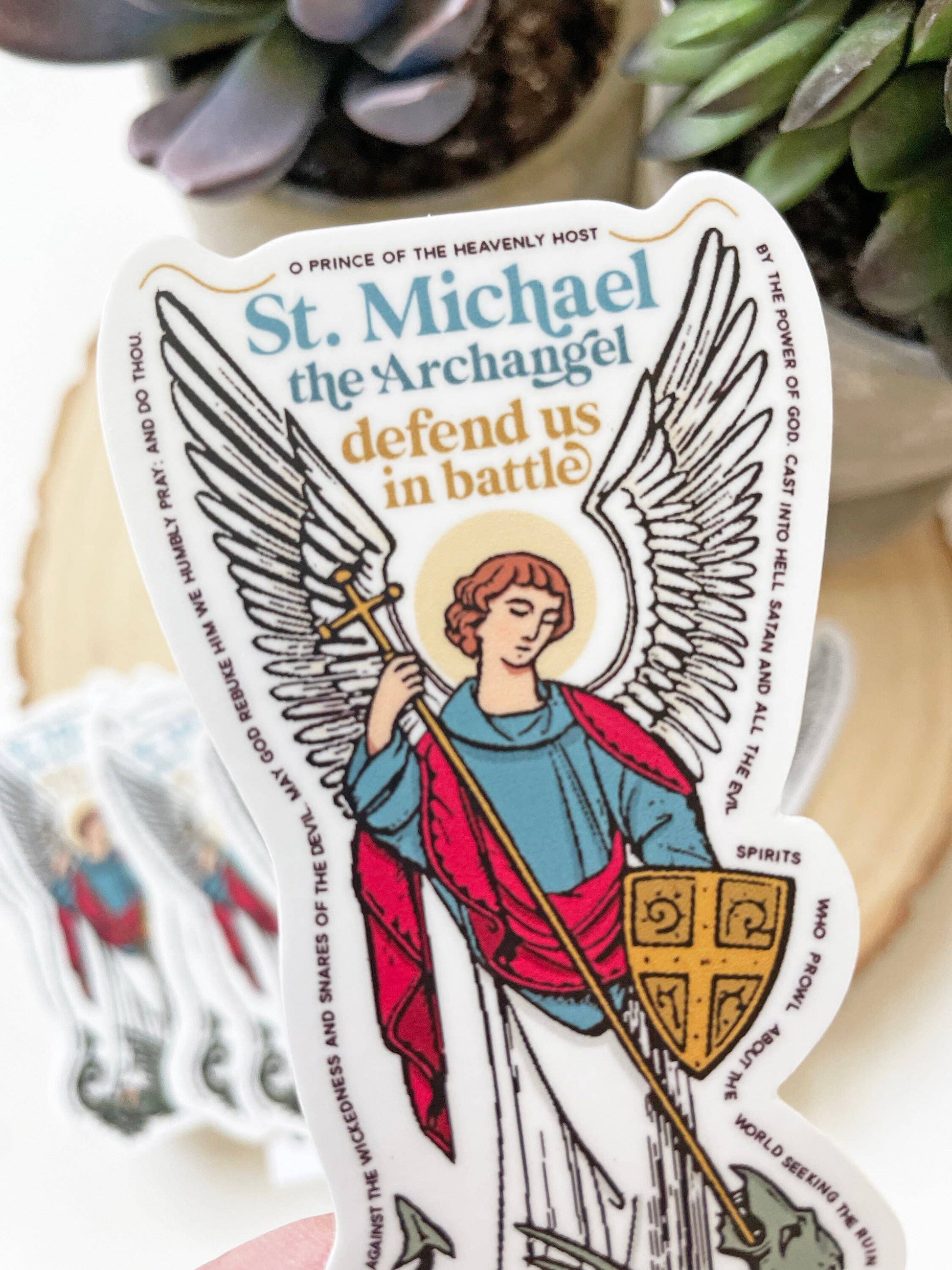 Sticker of St. Michael the Archangel with text and wings, placed on a surface with plants in the background.